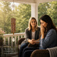 Two women sitting on a porch next to a hanging memorial wind chime. One woman appears distressed, covering her face with her hand, while the other sits beside her with a concerned expression.